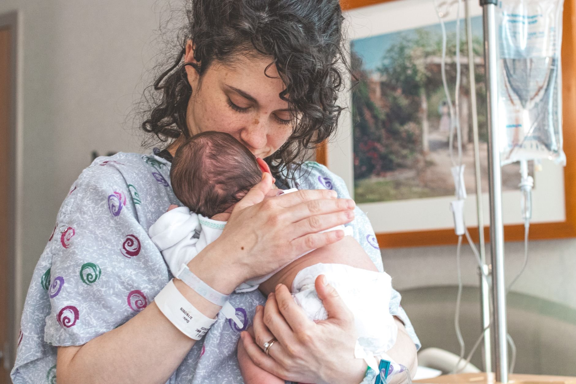 Mother in a hospital gown with curly dark hair kissing a newborn wrapped in a white blanket, with an IV pole beside her