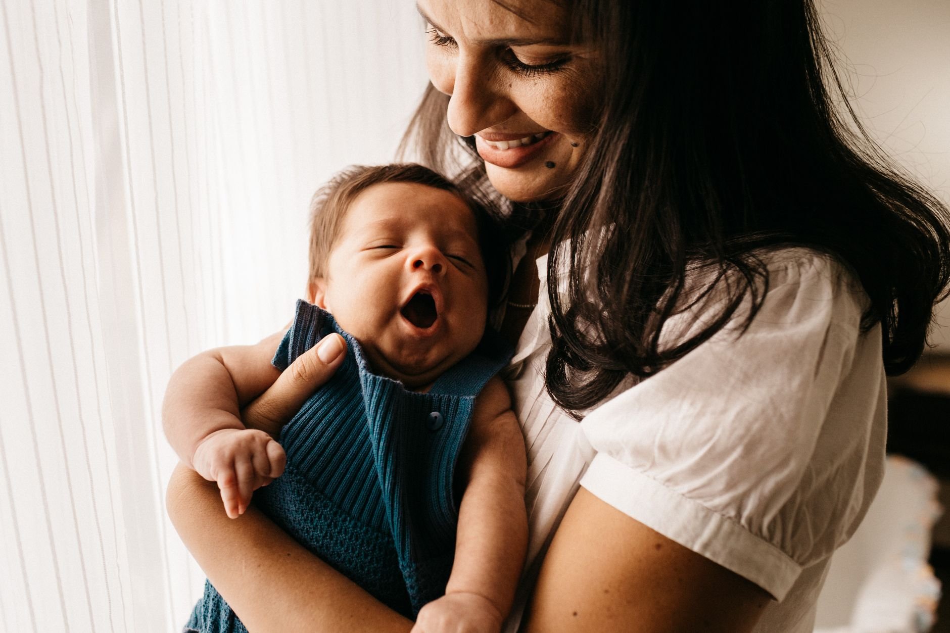 Smiling mother with long dark hair holding a yawning baby in a blue outfit near a sheer window