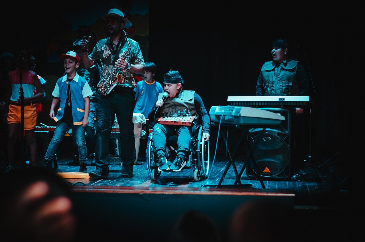 Performers on a dimly lit stage: a man in a hat playing saxophone, a seated performer in a wheelchair singing into a microphone, a keyboard player at right, and children at left