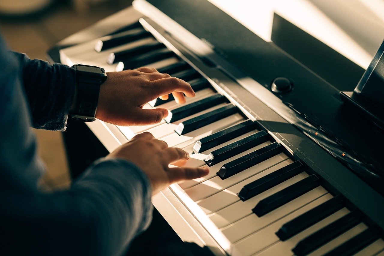Two hands playing a keyboard with black and white keys under warm light, one wrist wearing a watch