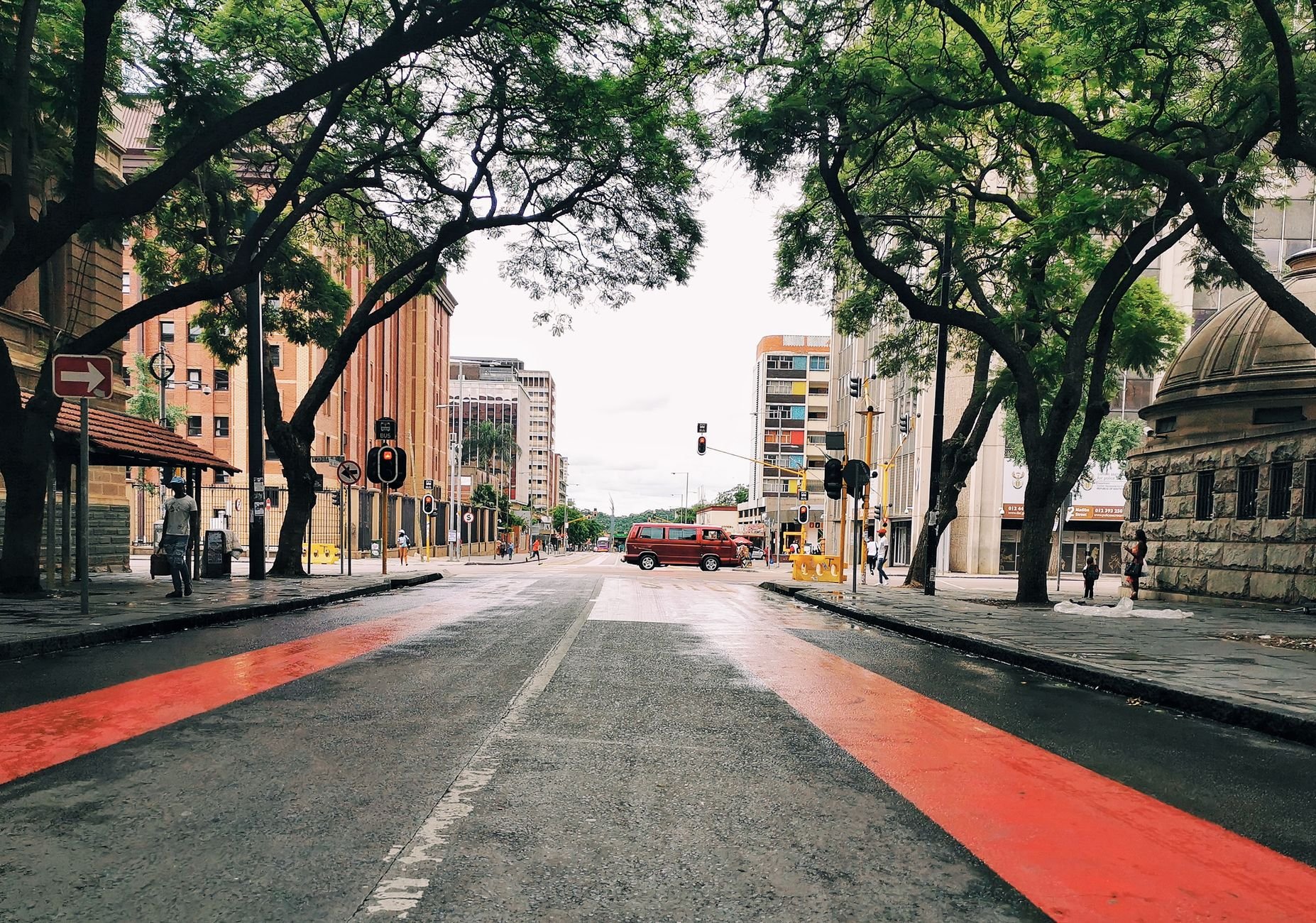 Empty city street in daylight with red-painted lanes, tall trees on both sides, multi-storey buildings, and a red van in the distance