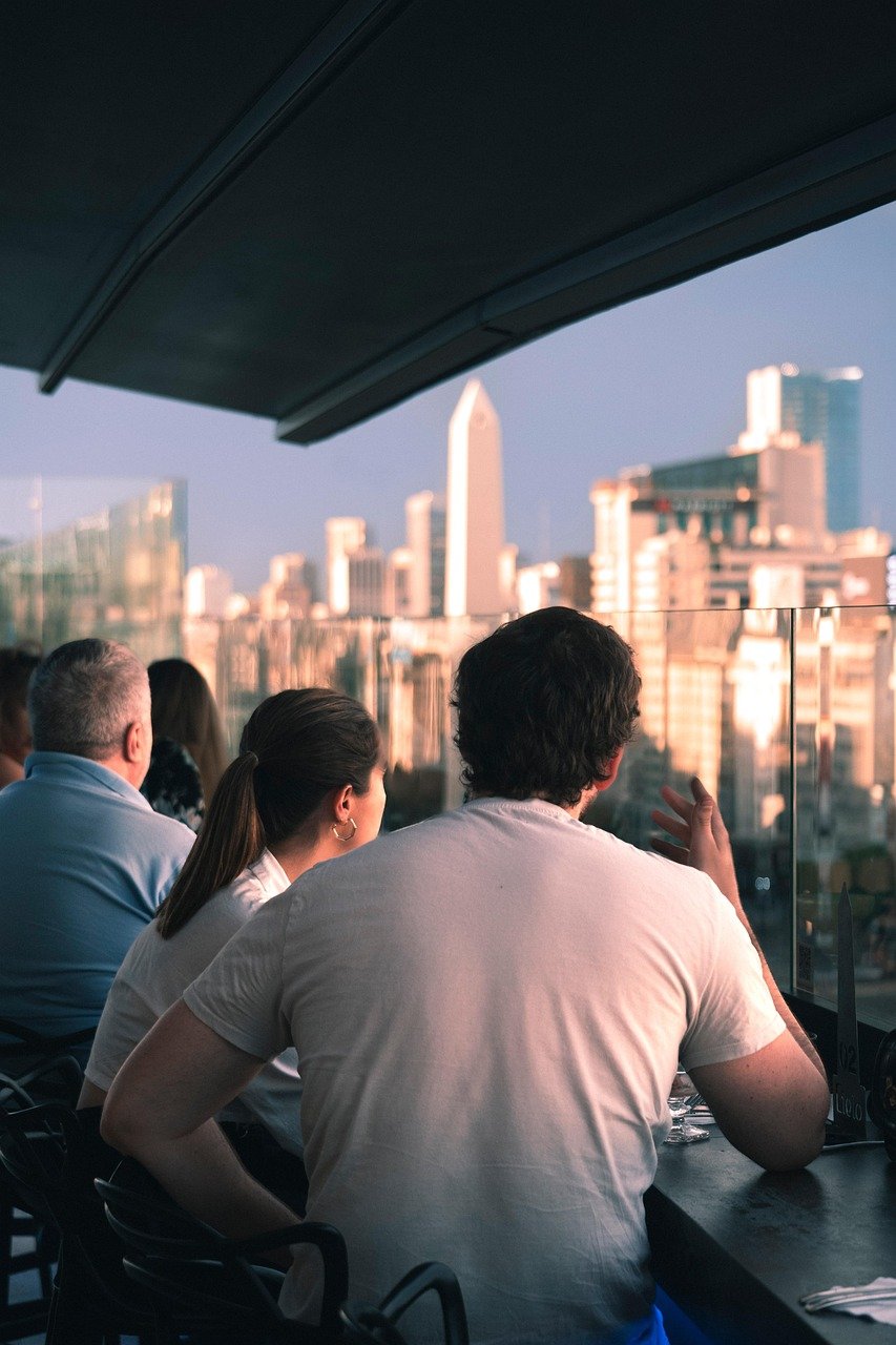 Couple seated at a rooftop bar with a city skyline behind a glass railing
