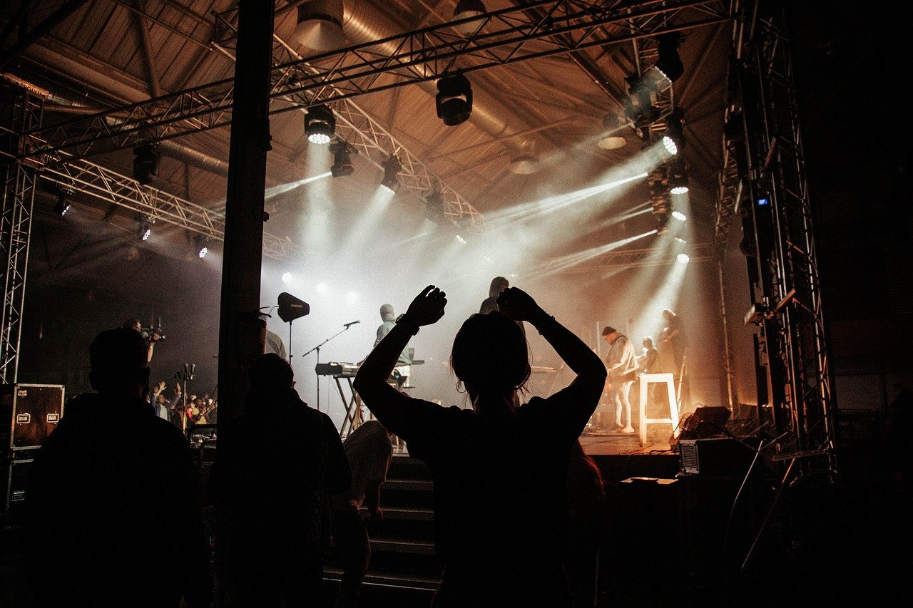 Silhouetted band on a lit stage with a crowd of people in the foreground, one with arms raised