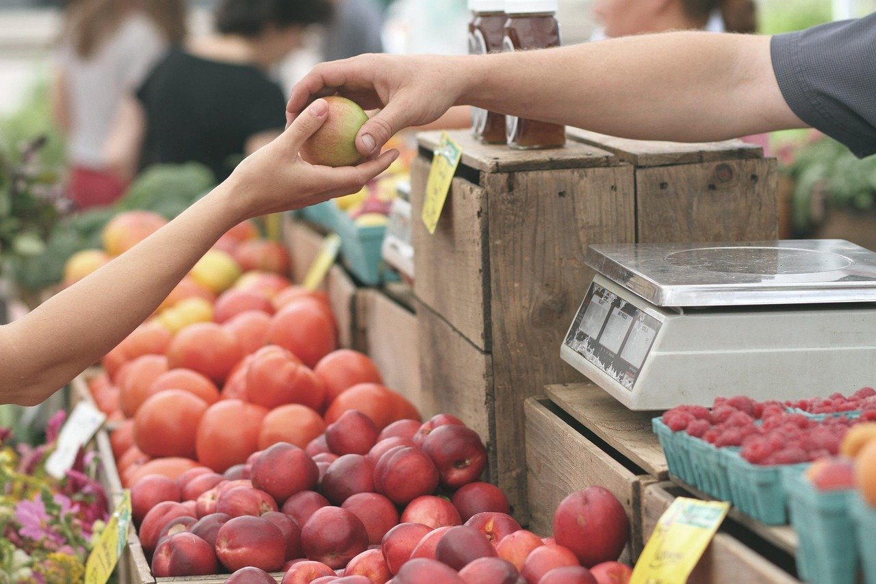 Hands exchanging a green apple over a wooden crate of peaches at an outdoor fruit stall