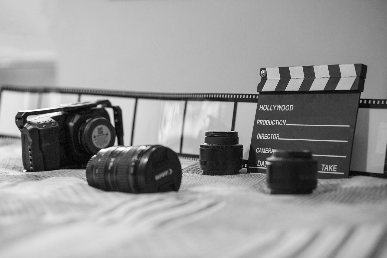 Black-and-white still life of a DSLR camera, a camera lens lying in front, and a clapperboard reading HOLLYWOOD PRODUCTION, DIRECTOR, CAMERA, and TAKE, with a film strip behind