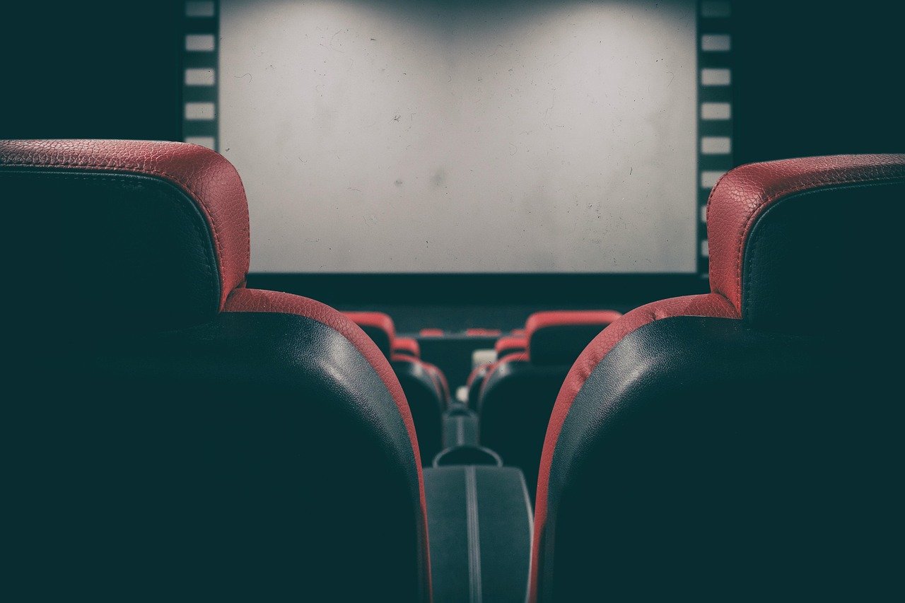 Rows of dark seats with red trim facing a blank white cinema screen in a dim theater
