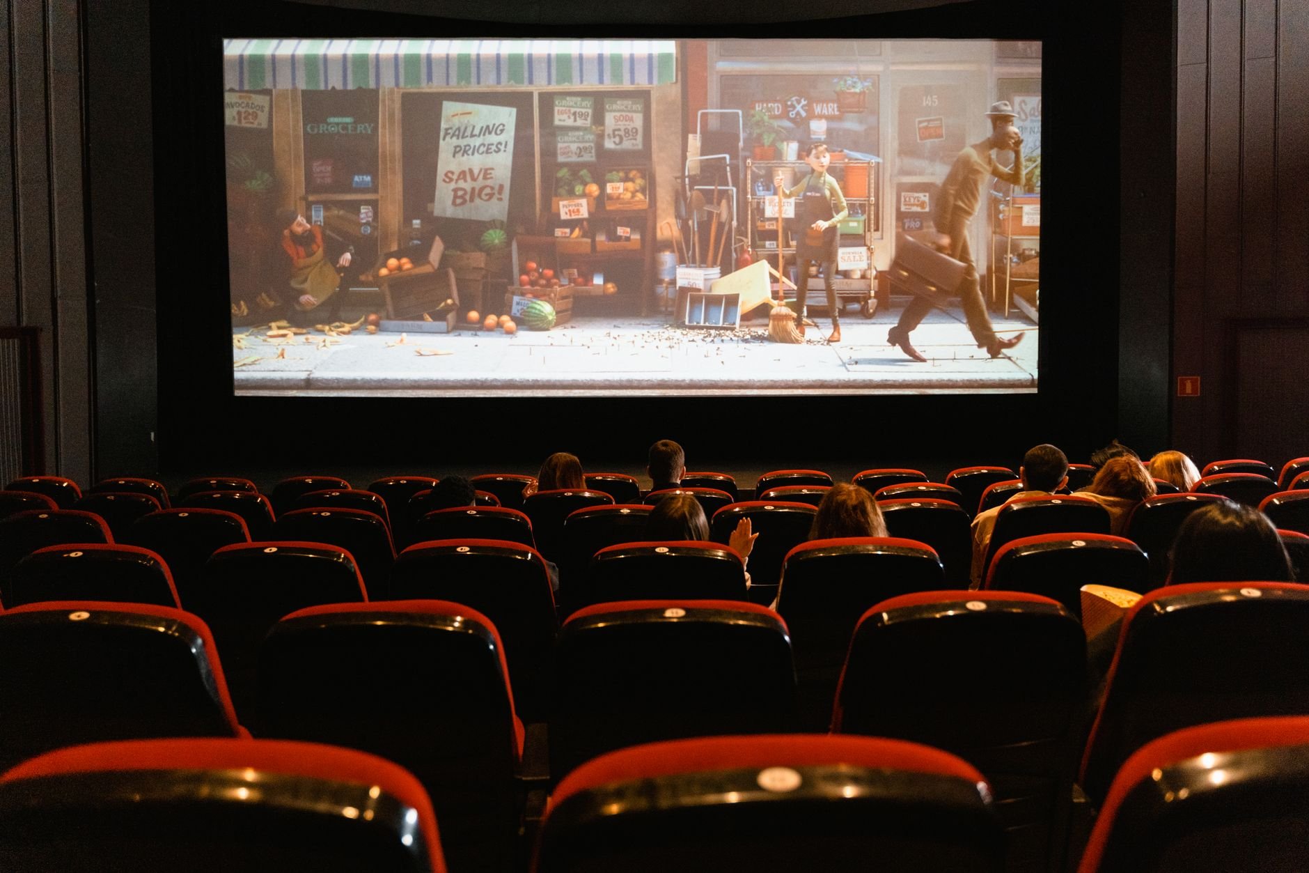 Trailer playing on a cinema screen above rows of red-seated theater chairs, with audience heads visible in the foreground