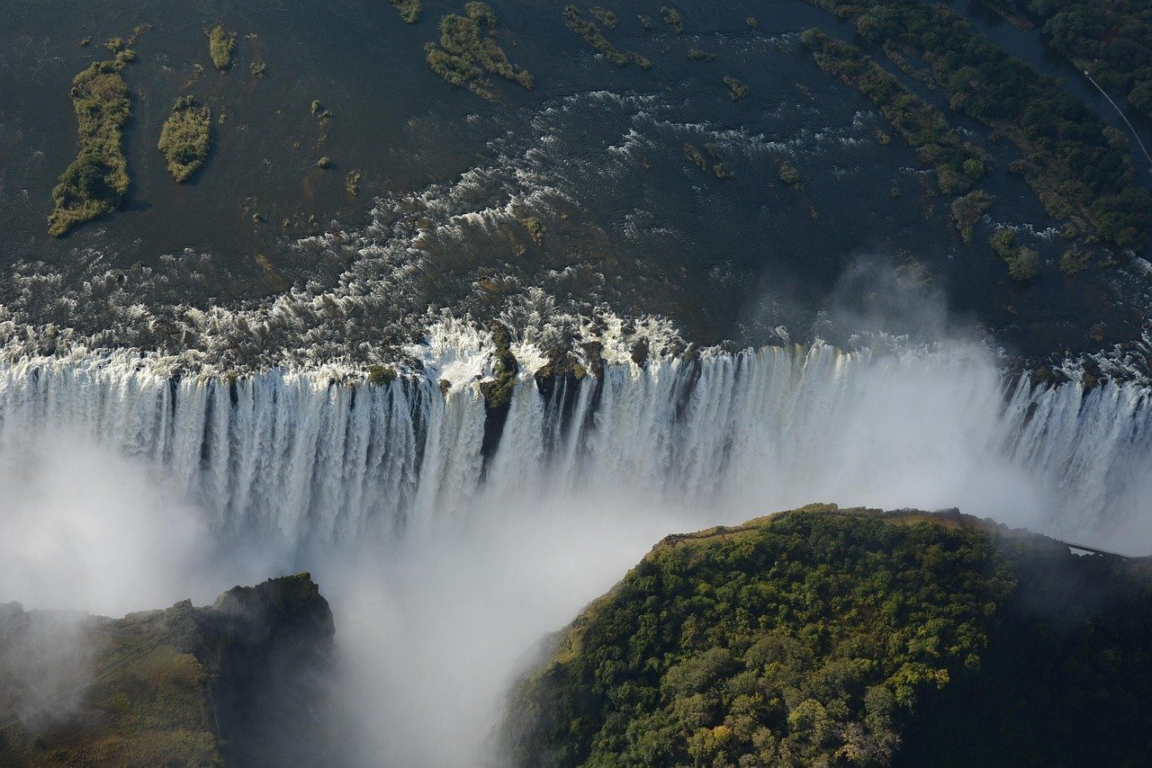 Aerial view of a long curtain of Victoria Falls with mist rising, dark green river land above and forested cliffs below