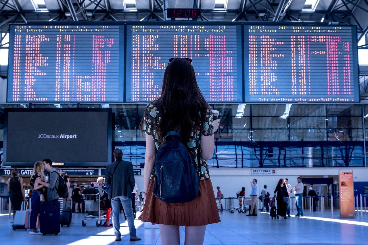Traveler with a backpack facing a large airport departures board showing flight information, with luggage carts and passengers in the terminal