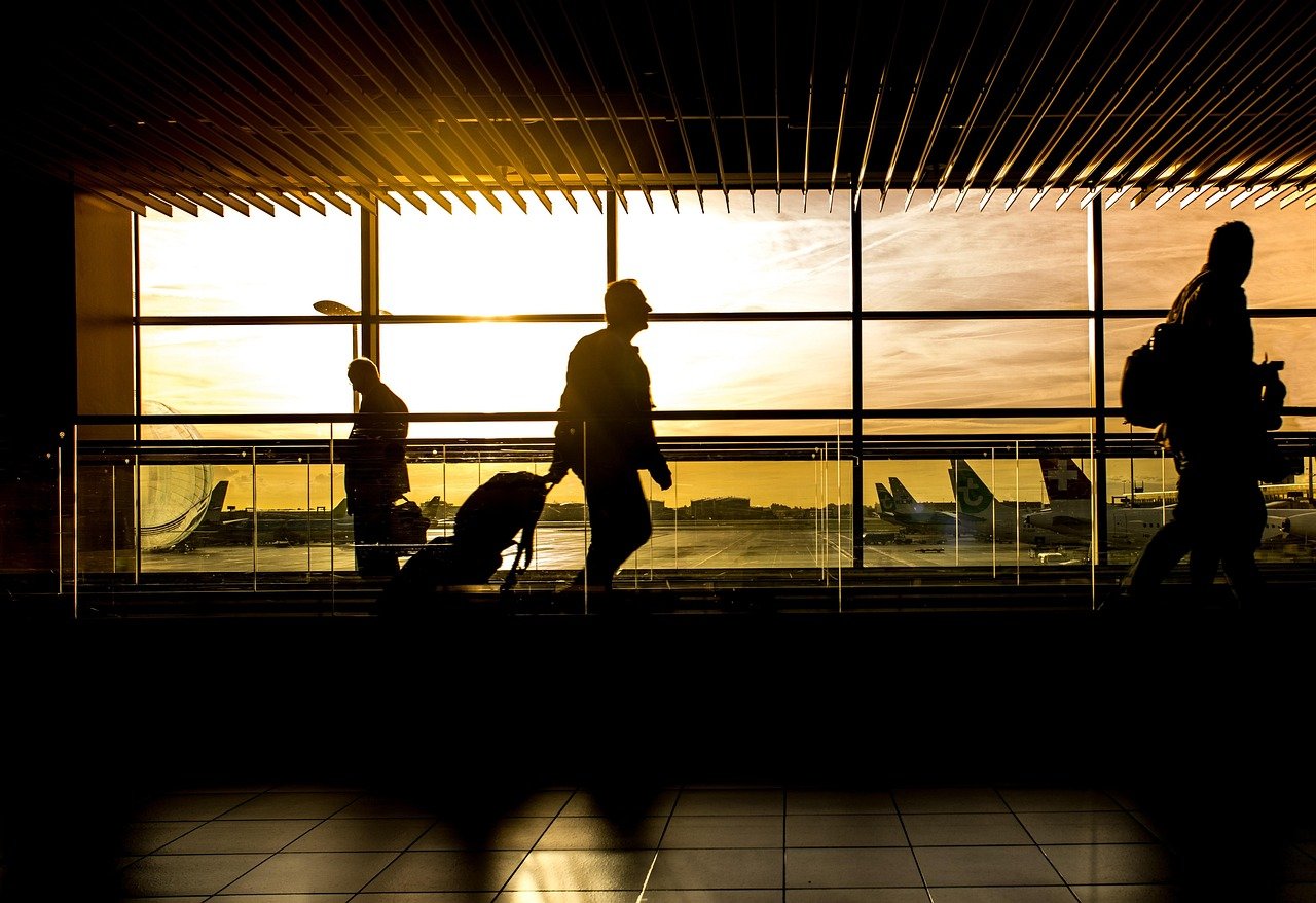Silhouetted travelers with rolling luggage walking past a large window at sunset, with aircraft parked on the apron outside