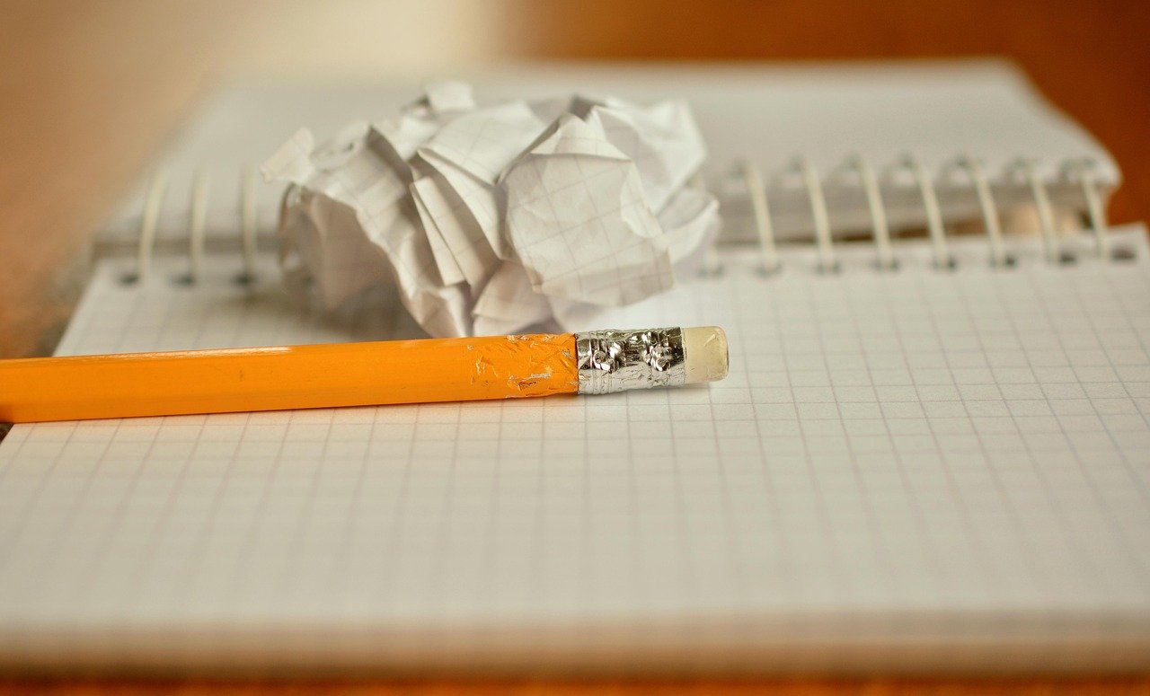 Yellow pencil with a worn eraser lying across a grid-ruled notebook page, with a crumpled paper ball behind it
