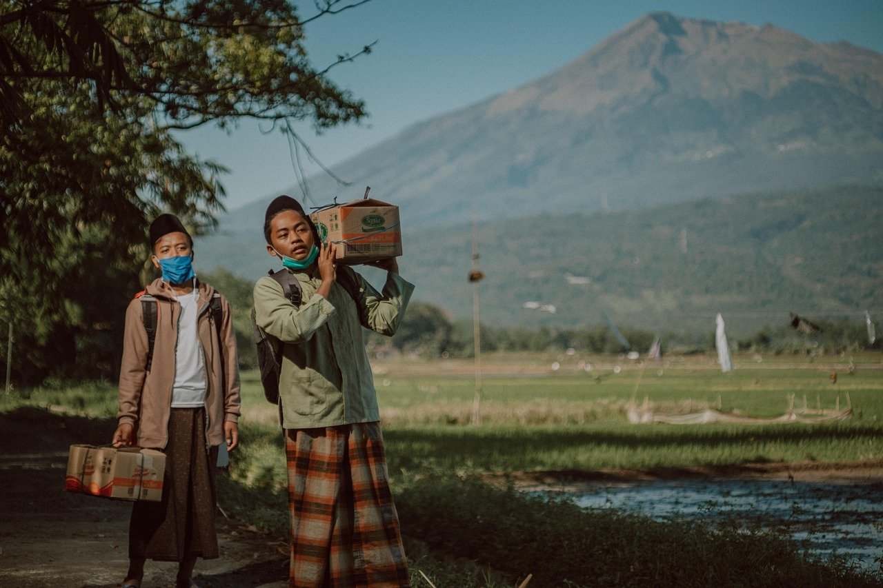 Two young people walk along a dirt path past a rice field, one carrying a cardboard box on his shoulder and the other holding a box, with a mountain in the background