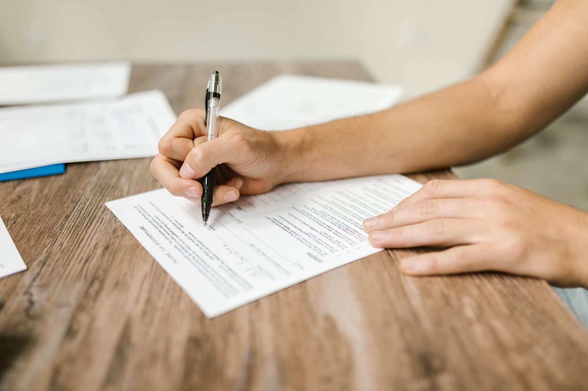 A hand holding a pen over a printed form with lines of small text, on a wooden table