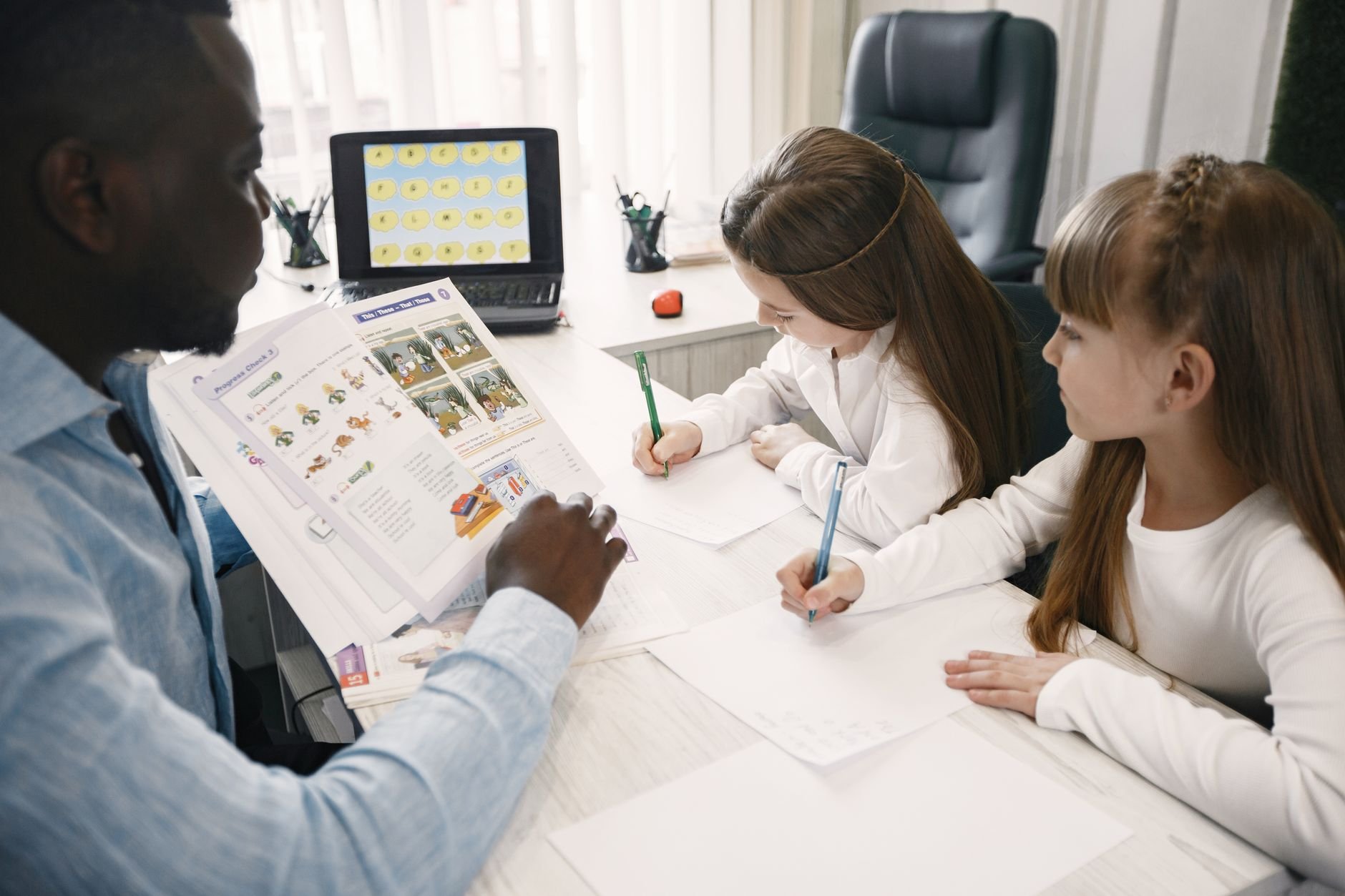 An adult man points at a printed sheet with pictures of plants and animals while two girls write on paper at a desk with a laptop and pencil cups nearby