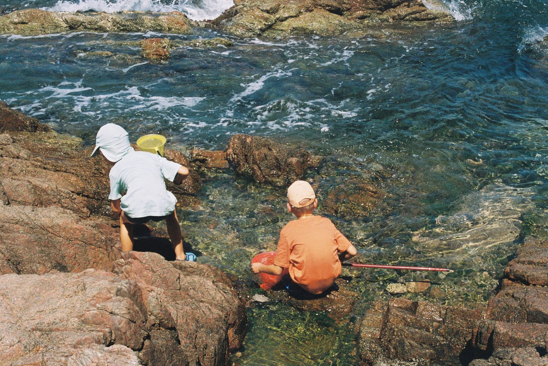 Two children on a rocky shore beside clear green-blue water: one in a white sun hat and white shirt stands on a rock holding a yellow net, while the other, in an orange shirt, crouches at the water's edge holding a red container and a long pole.