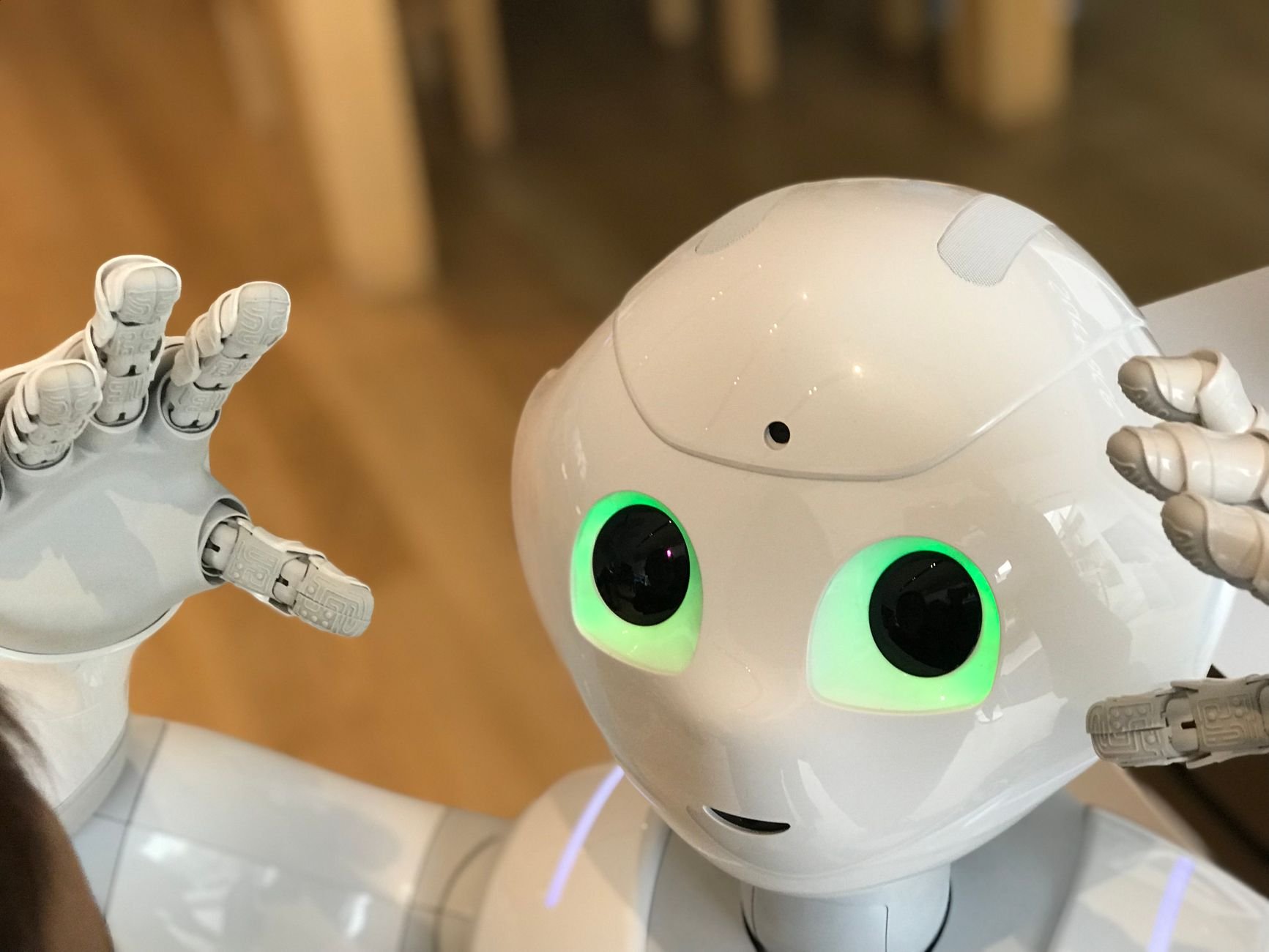 White humanoid robot with glowing green eyes and a raised hand in the foreground, with a blurred indoor background