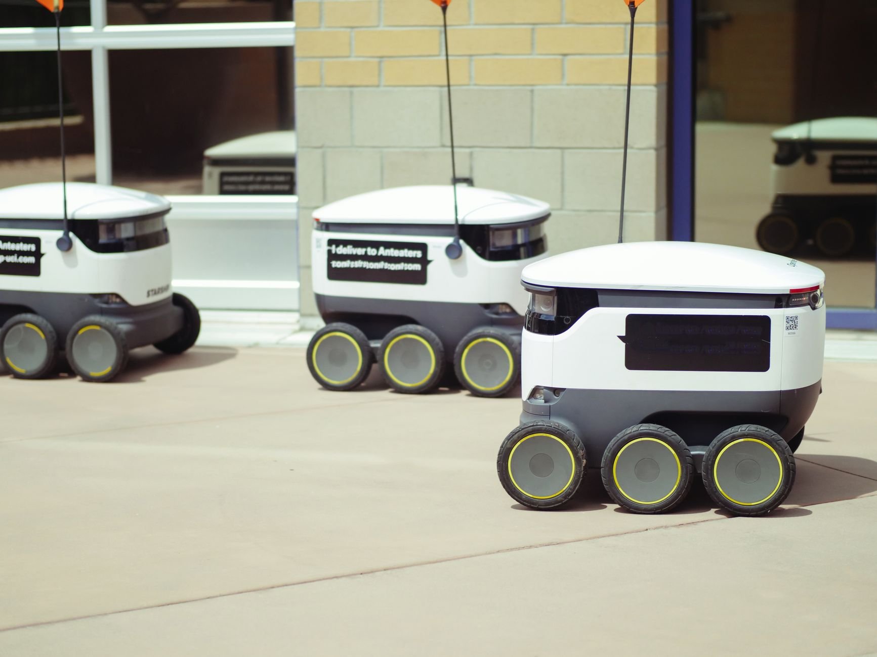 Three white wheeled delivery robots with six wheels and yellow hubs parked on a paved courtyard in front of a brick building