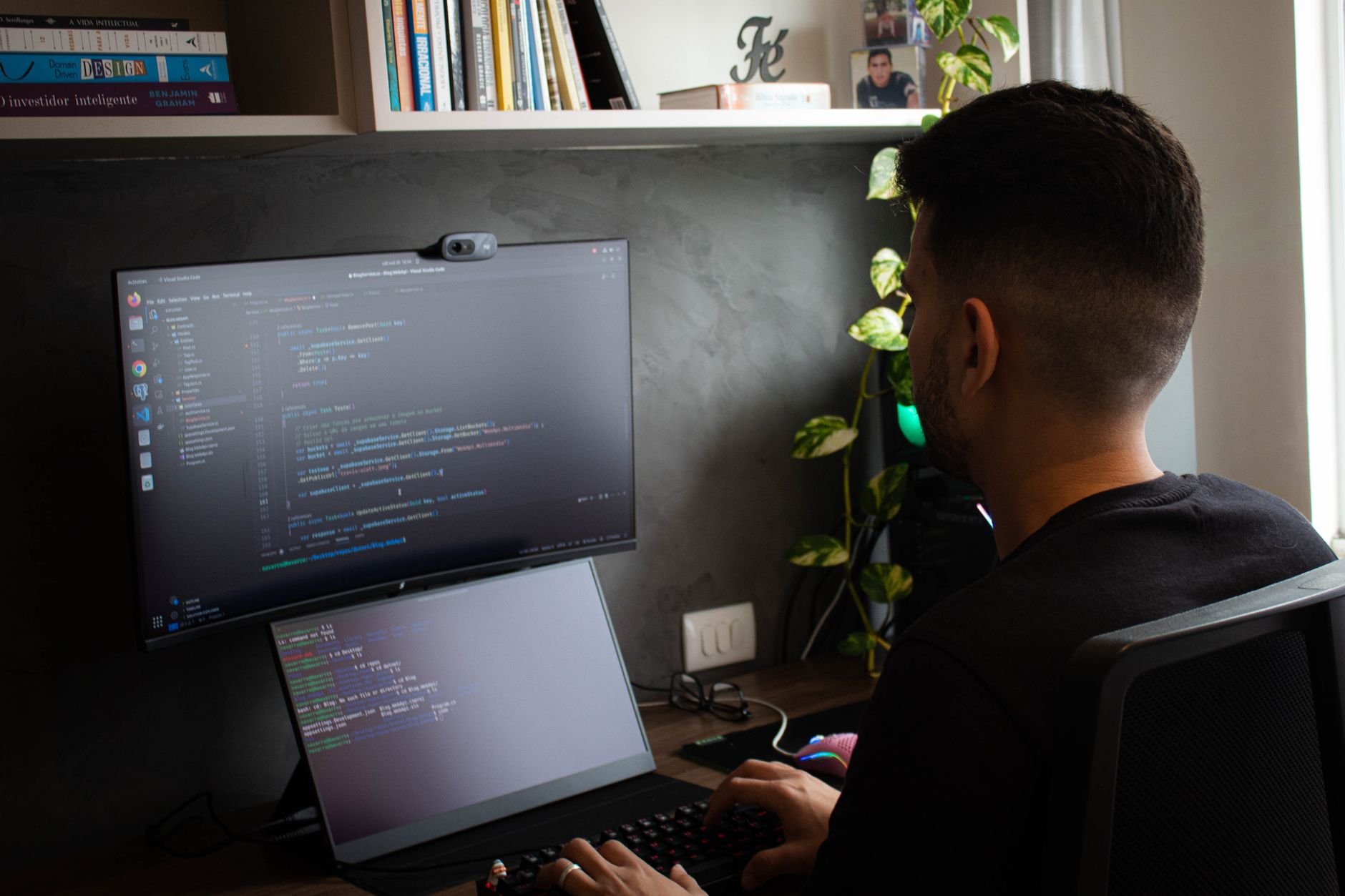 Man seated at a desk working on code across a monitor with a webcam mounted on top and a laptop showing code below