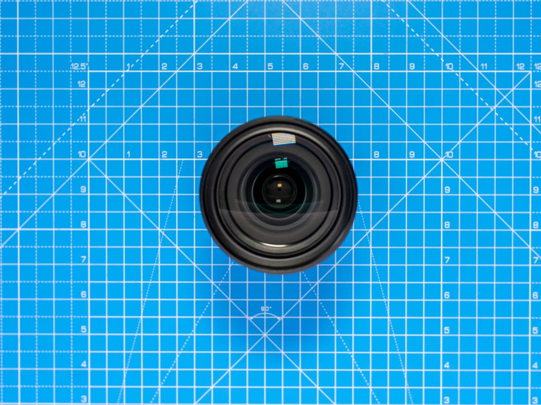 Black camera lens viewed from above, resting on a blue cutting mat printed with a grid