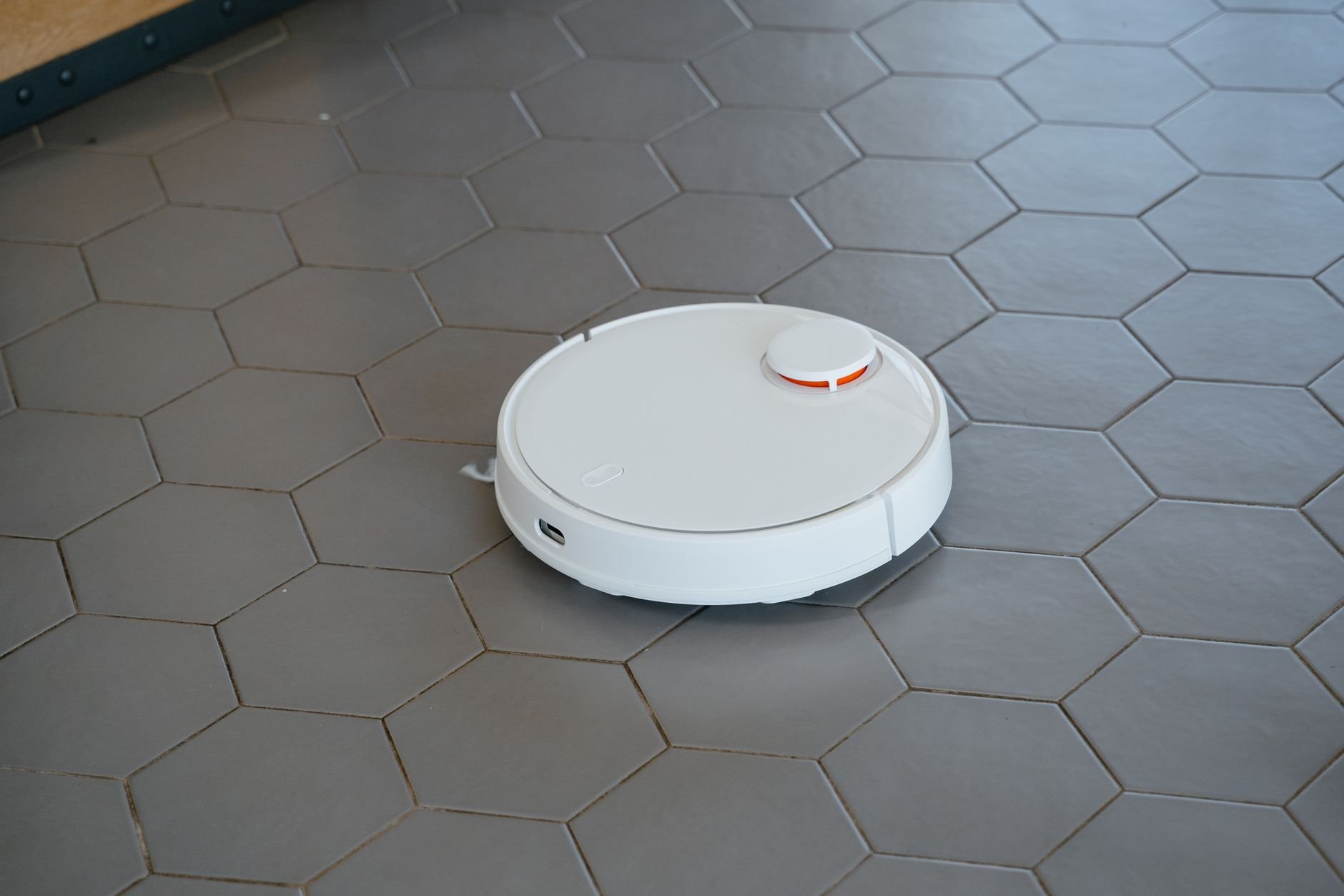 White round robot vacuum with a red-orange sensor on its top, moving across grey hexagonal floor tiles