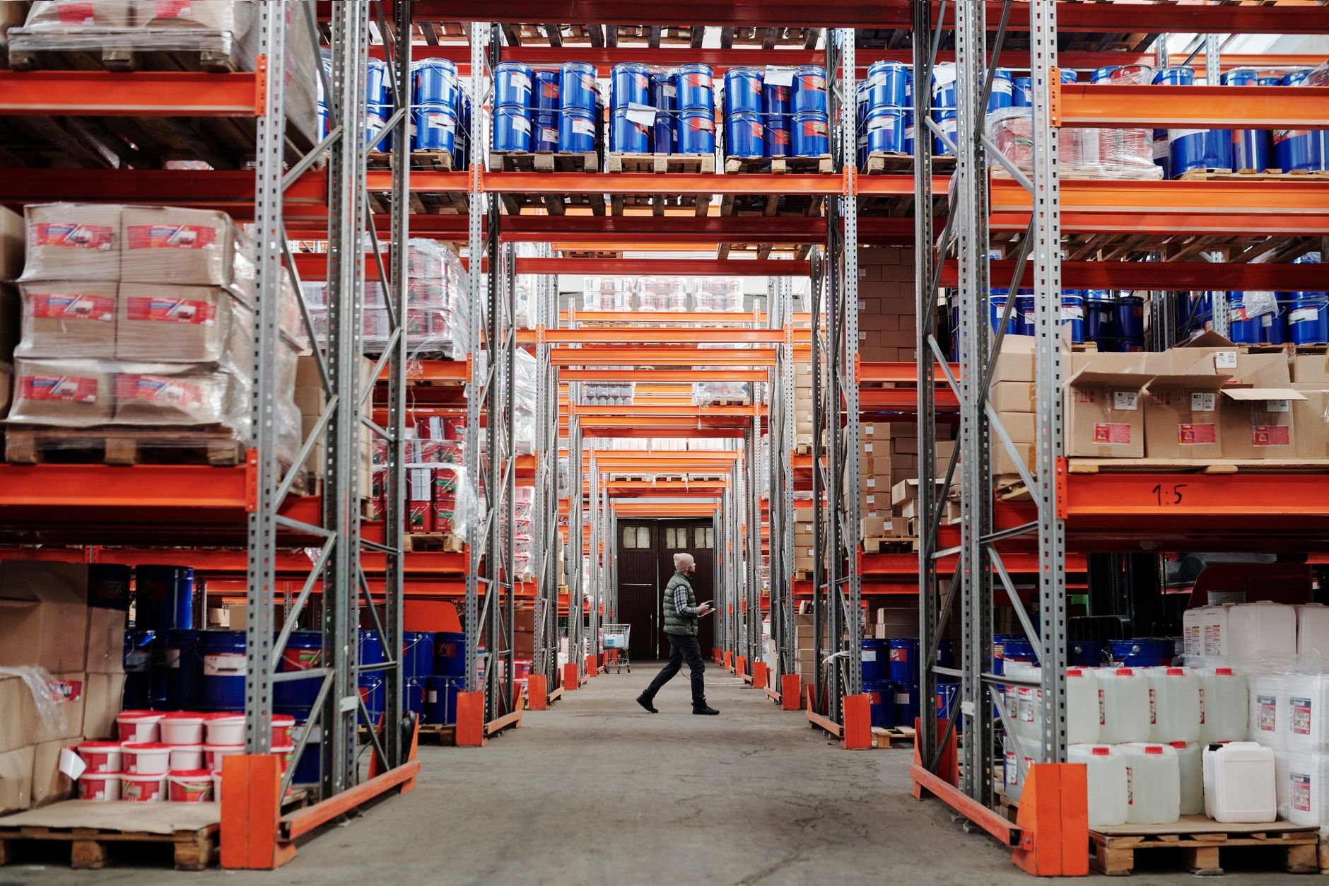 Warehouse aisle lined with tall orange and grey shelving holding boxes and blue drums, with one worker walking down the central aisle