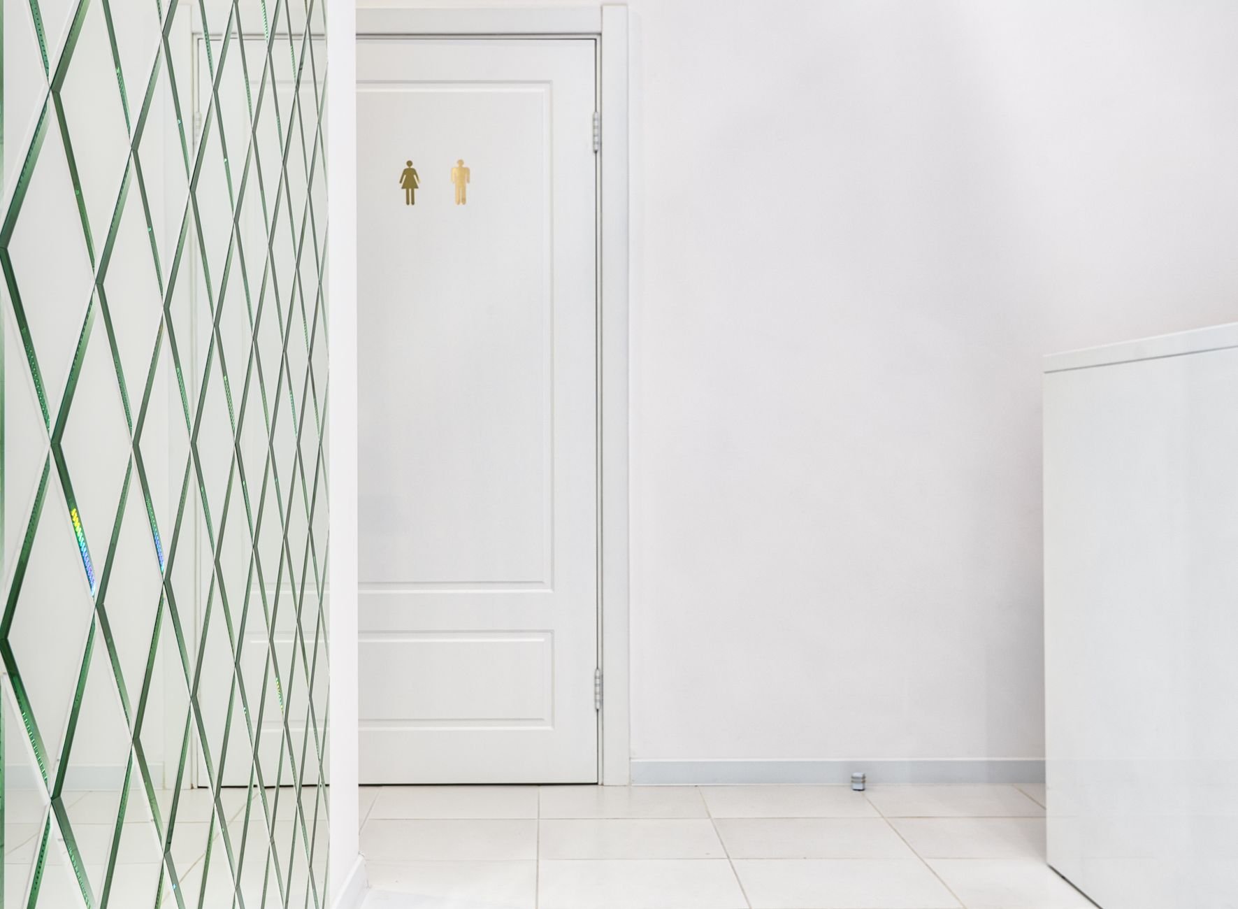 Indoor corridor with a white panelled door marked with restroom symbols, a patterned glass partition, and a plain white wall