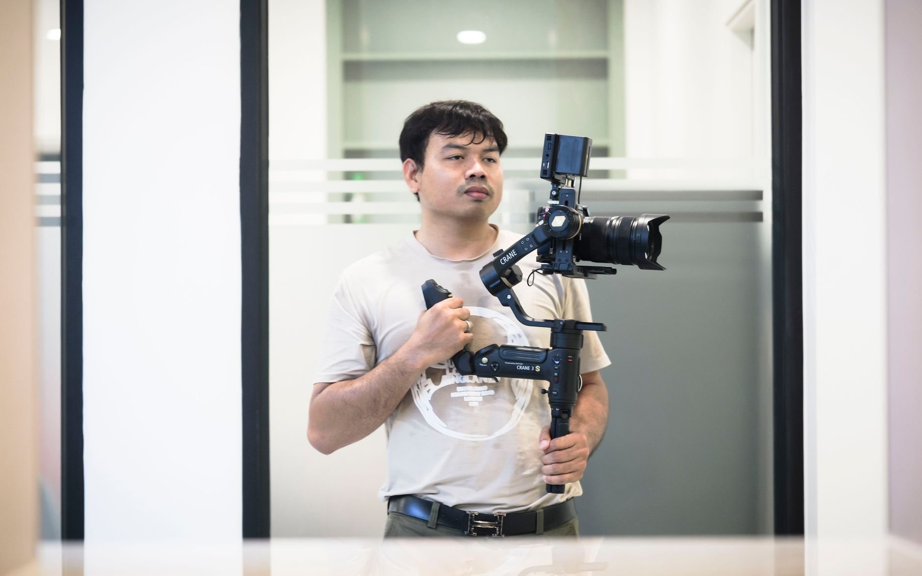 Man holding a handheld gimbal with a camera mounted on it, standing in an indoor office corridor