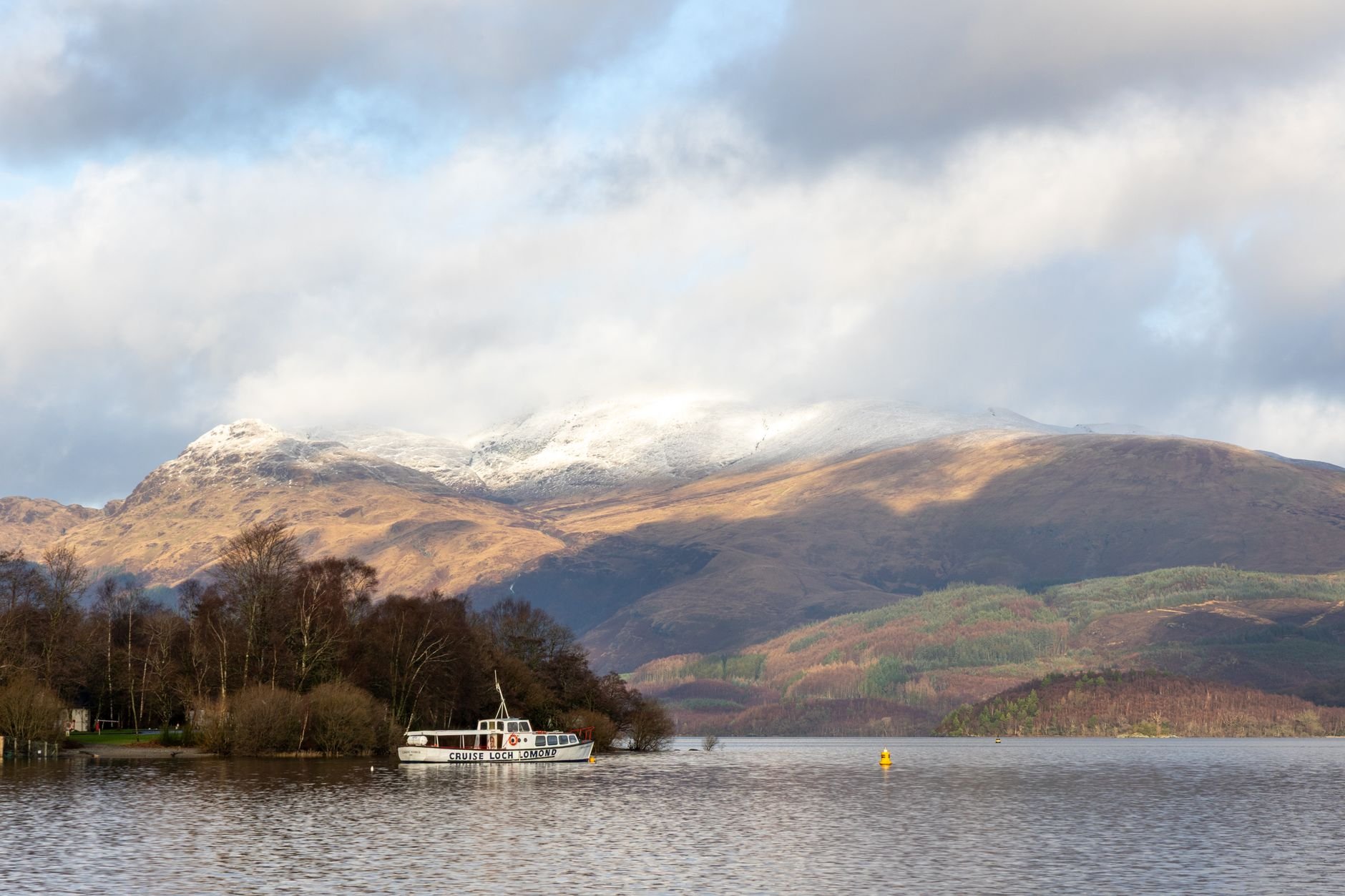 A white passenger boat moored on calm water in front of a wooded shore, with snow-dusted mountains and brown hillsides rising behind under broken cloud