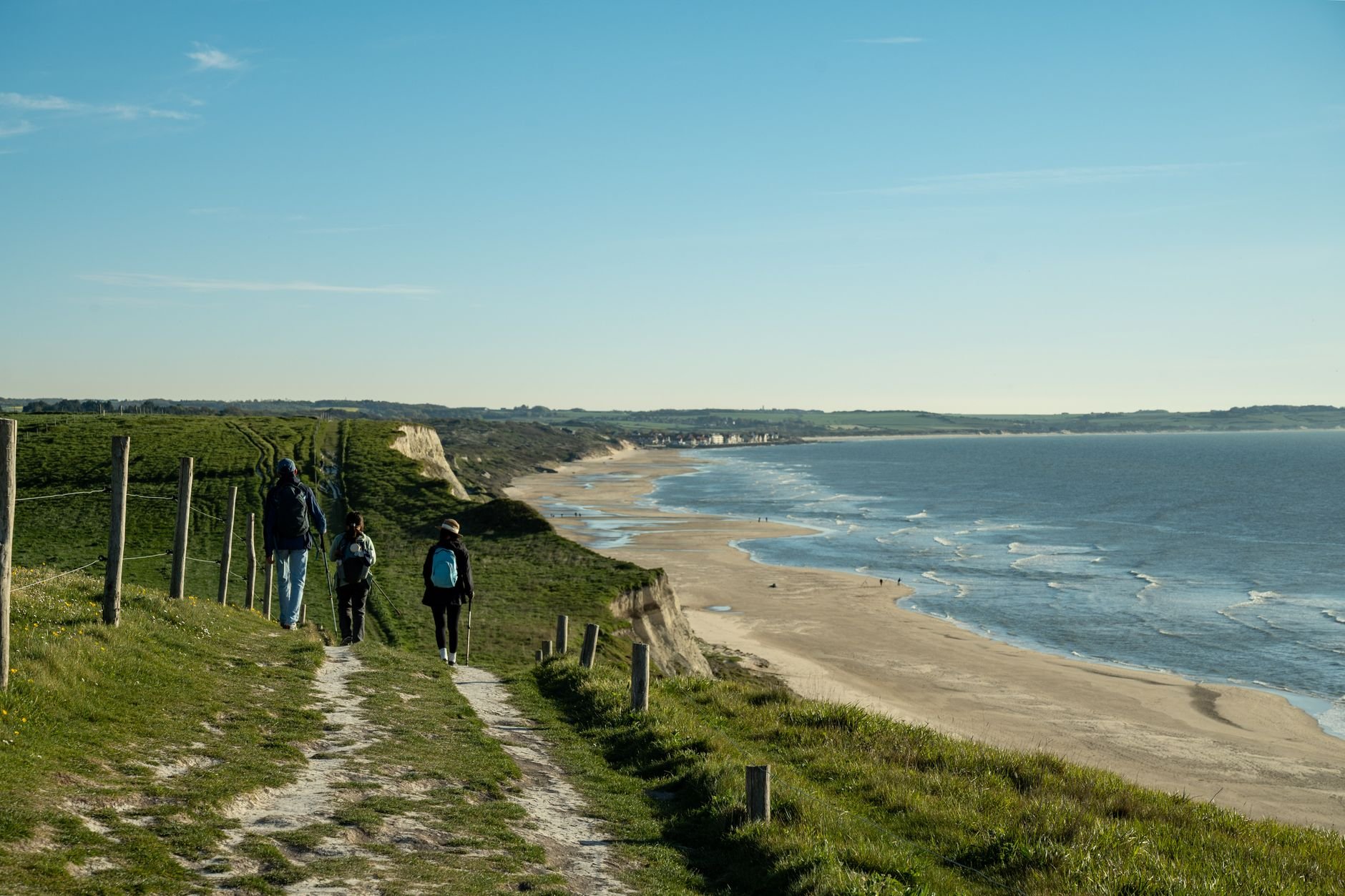 Three walkers with poles on a grassy coastal path above a sandy beach and the sea on a clear day