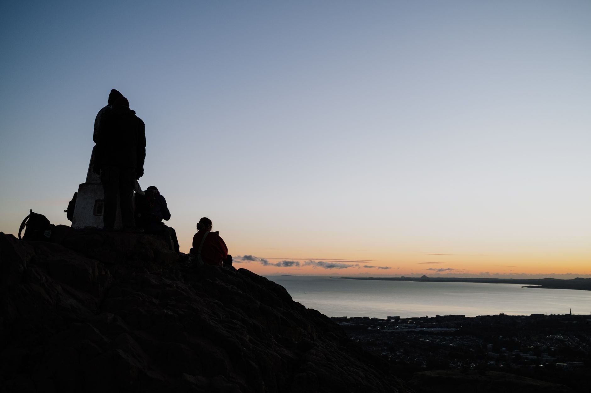 Silhouetted walkers on a rocky hilltop at sunset, with a city and water below