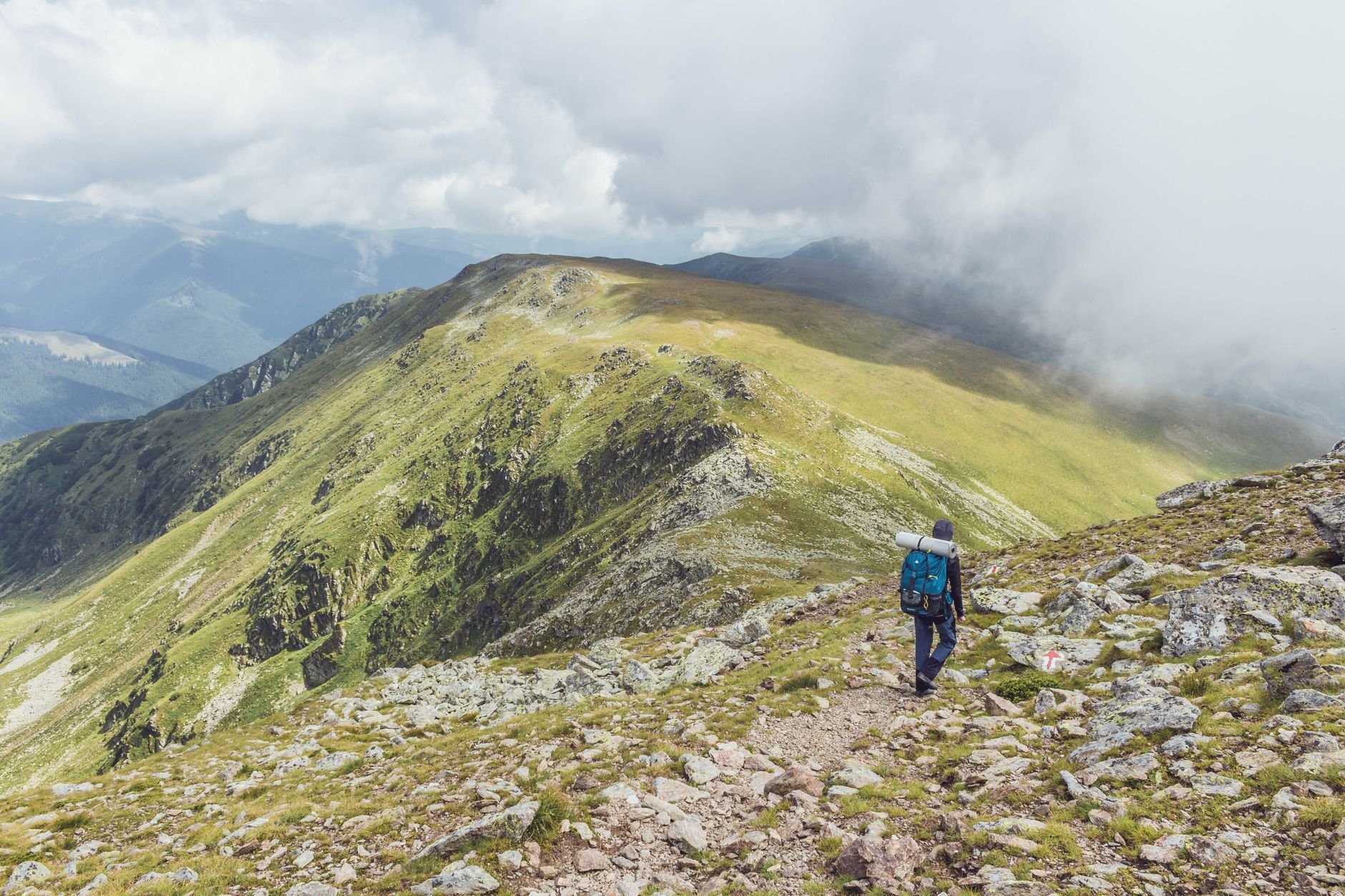 Hiker with a blue backpack walking along a grassy ridge path with cloud and distant mountains behind
