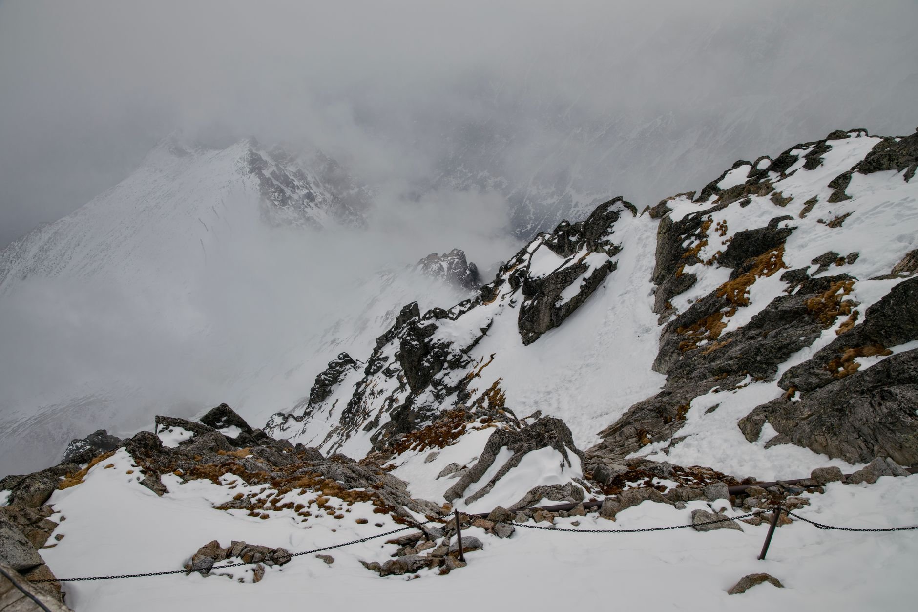 Snow-covered rocky ridge with a fixed wire cable running along it, with cloud and mist in the background