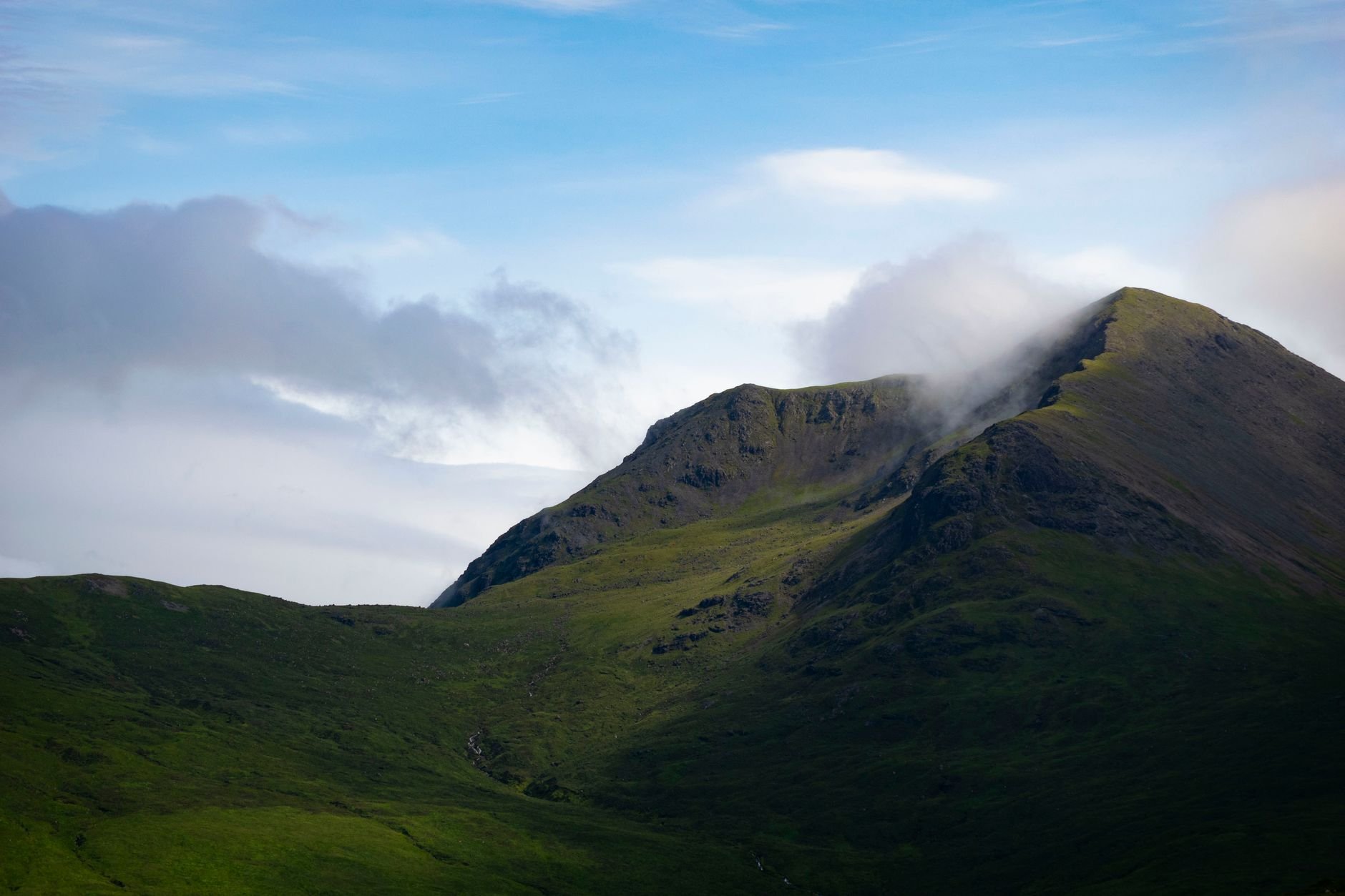 Green mountain summit with wisps of cloud clinging to the peak under a blue sky with scattered clouds