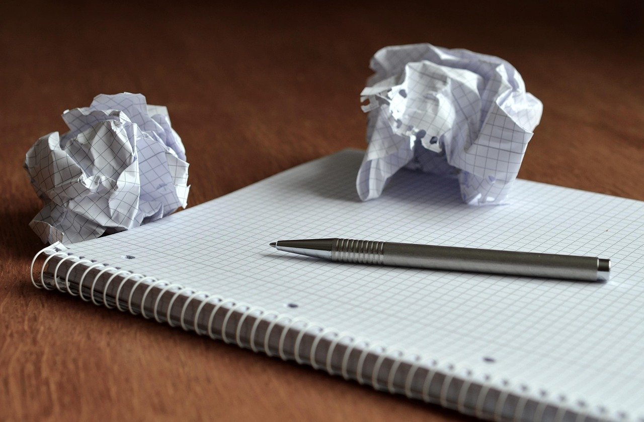 Silver pen resting on an open grid notebook beside two crumpled paper balls on a wooden table