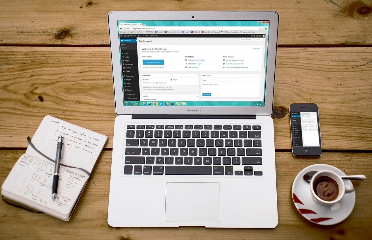 Silver laptop showing a WordPress dashboard next to an open handwritten notebook, a pen, a smartphone, and a cup of coffee on a wooden desk