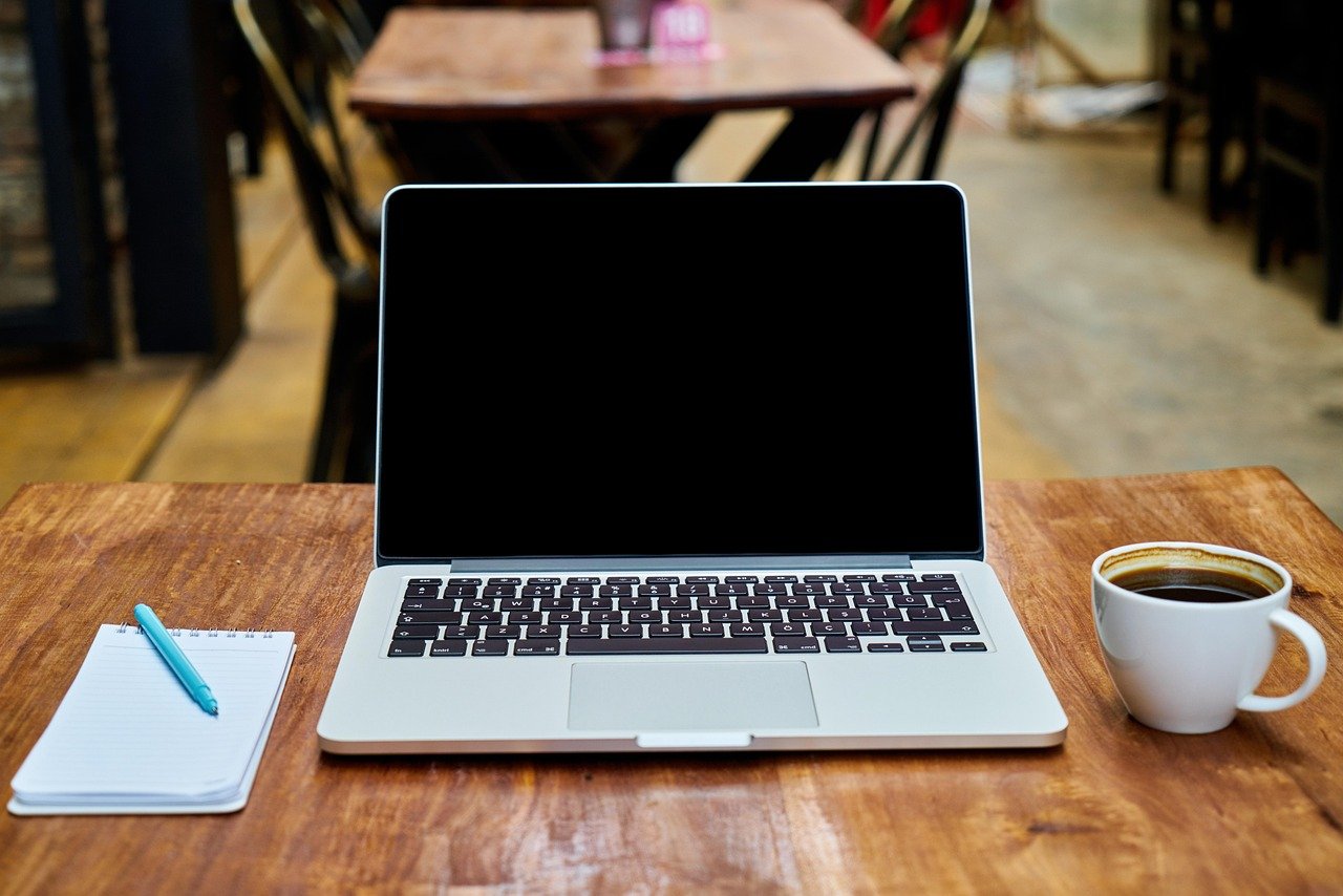 Open laptop with a blank black screen on a wooden table beside a spiral notepad with a teal pen and a white cup of coffee